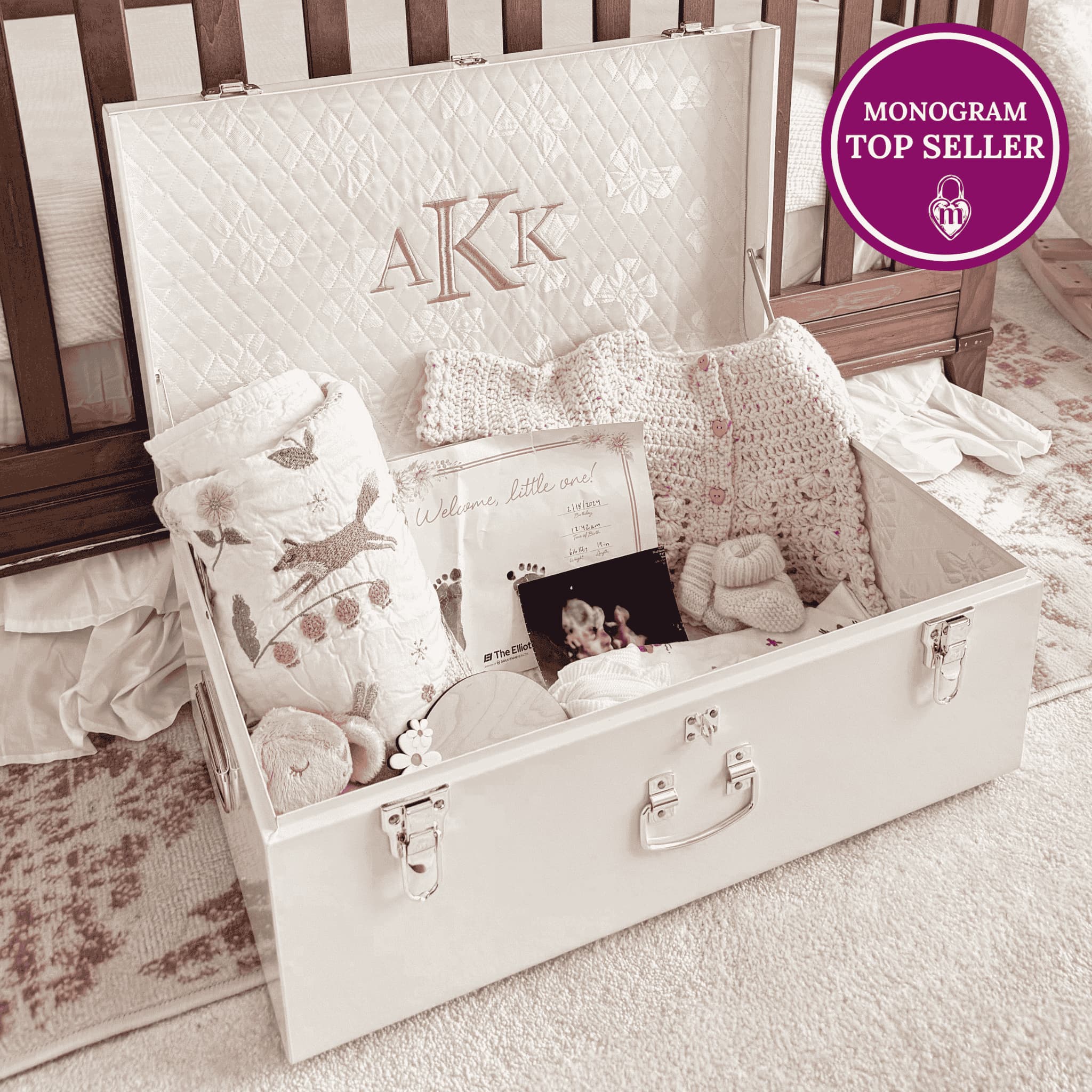 White baby trunk with baby items and monogrammed cover, sitting on a carpeted floor.