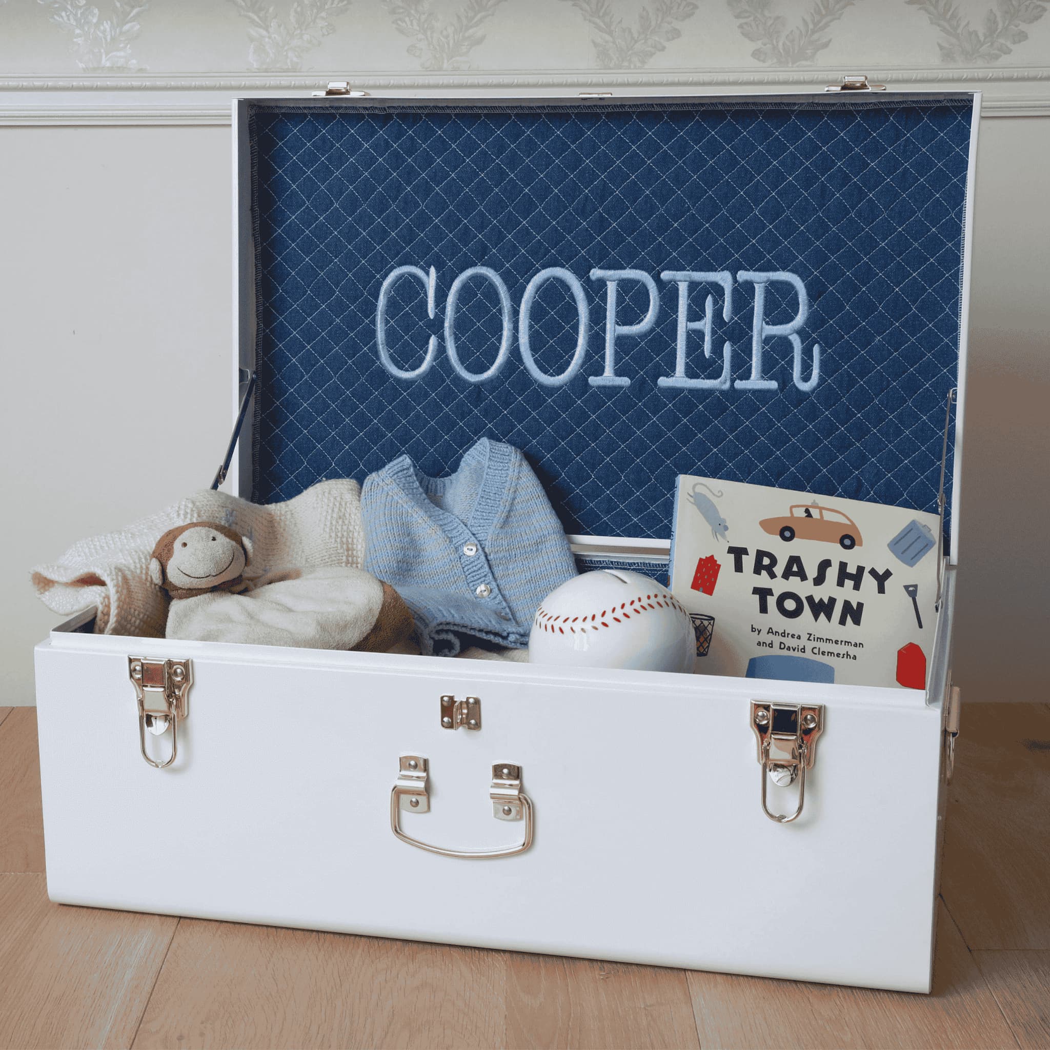 White toy chest with blue interior, containing a teddy bear, denim shirt, baseball, and book titled 'Trashy Town'.