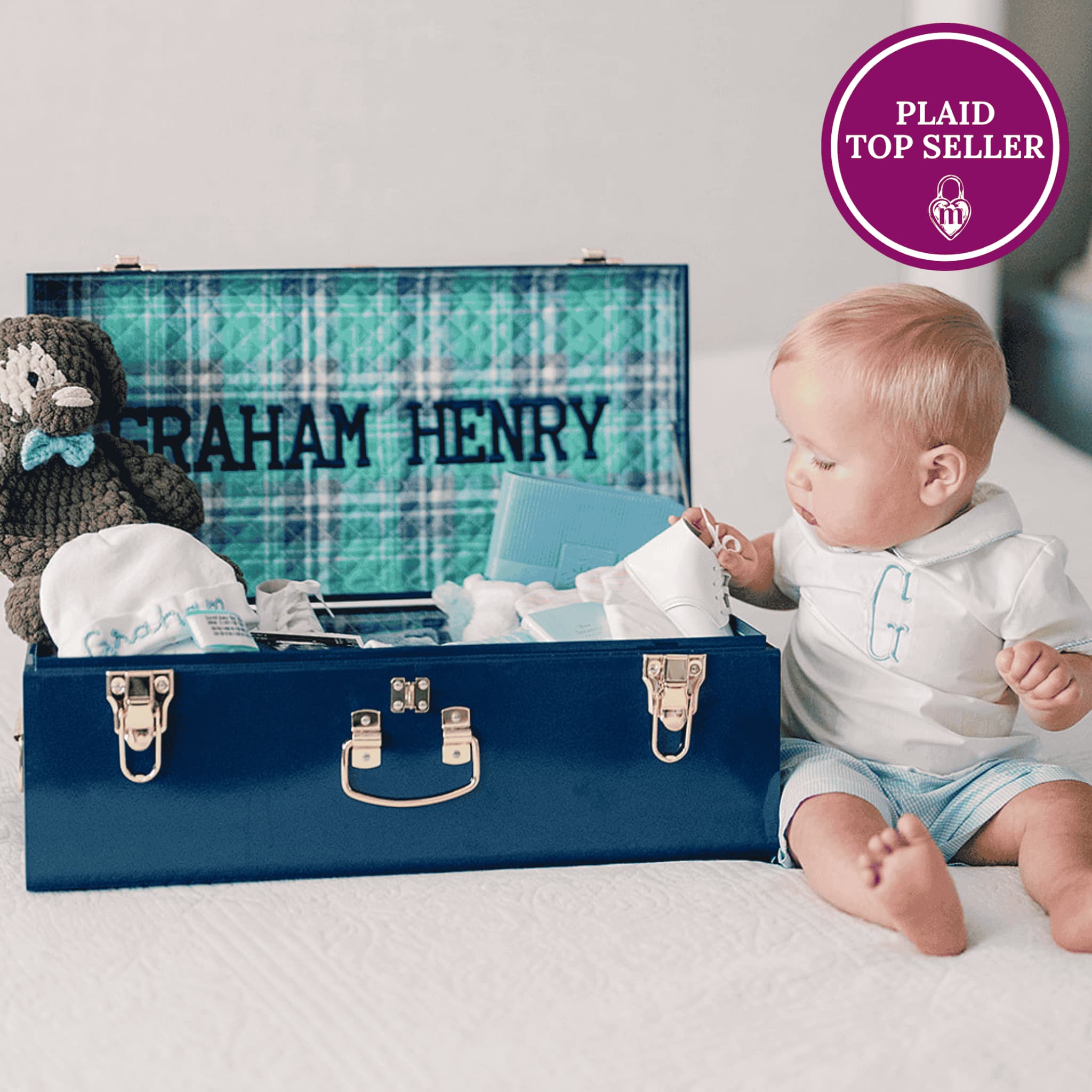 Baby with a keepsake trunk containing baby items and a teddy bear, with 'Plaid Top Seller' badge.