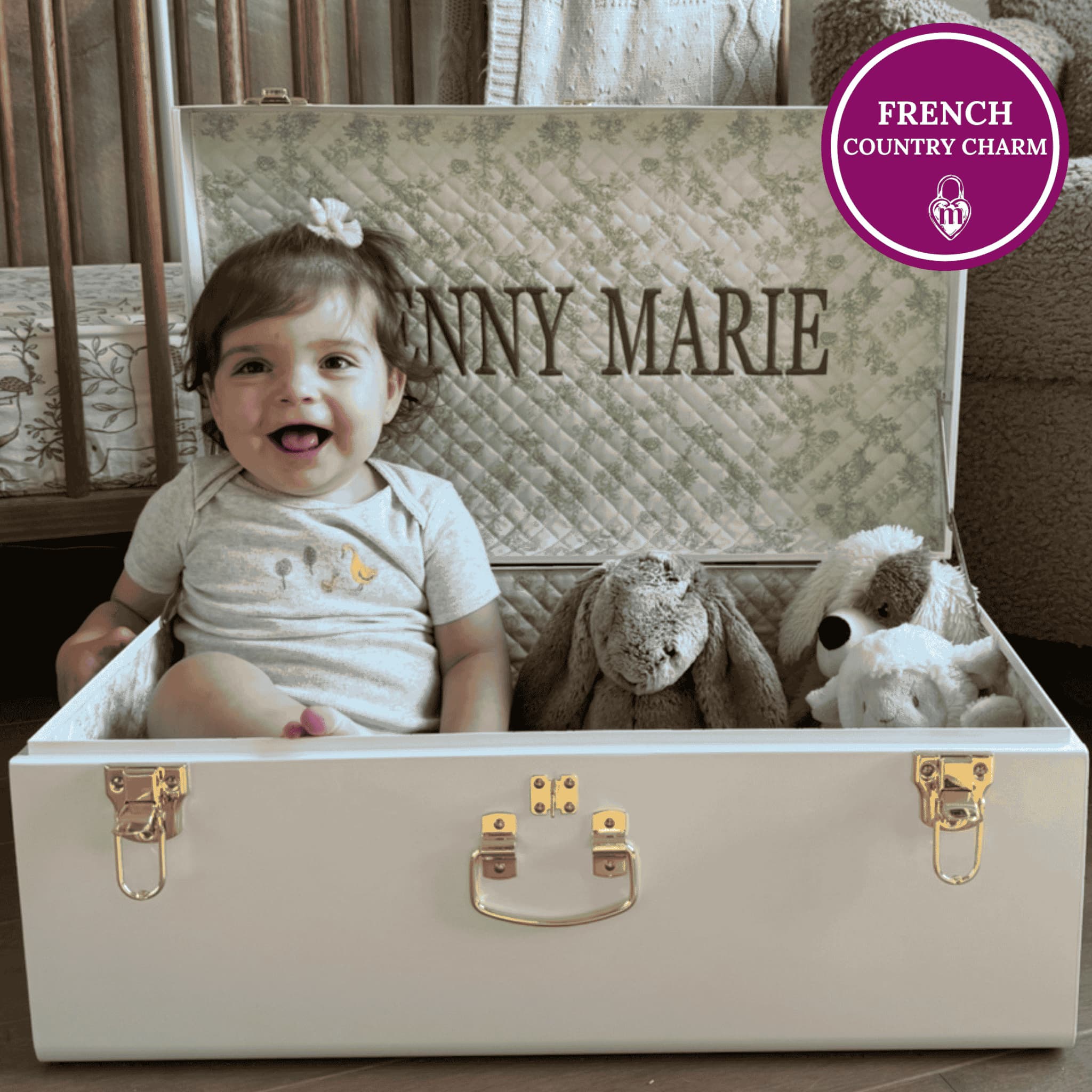 Child sitting in a keepsake trunk with embroidery on it, surrounded by stuffed animals.