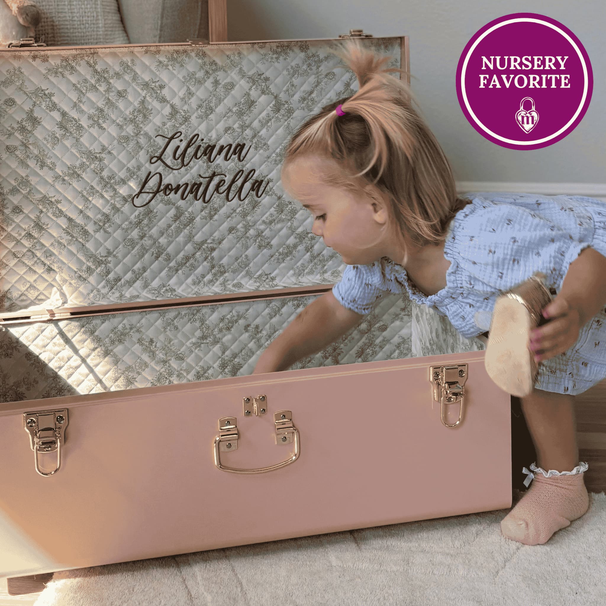 Child playing with a pink memory trunk with 'Liliana Donatella' embroidery, labeled 'Nursery Favorite'.